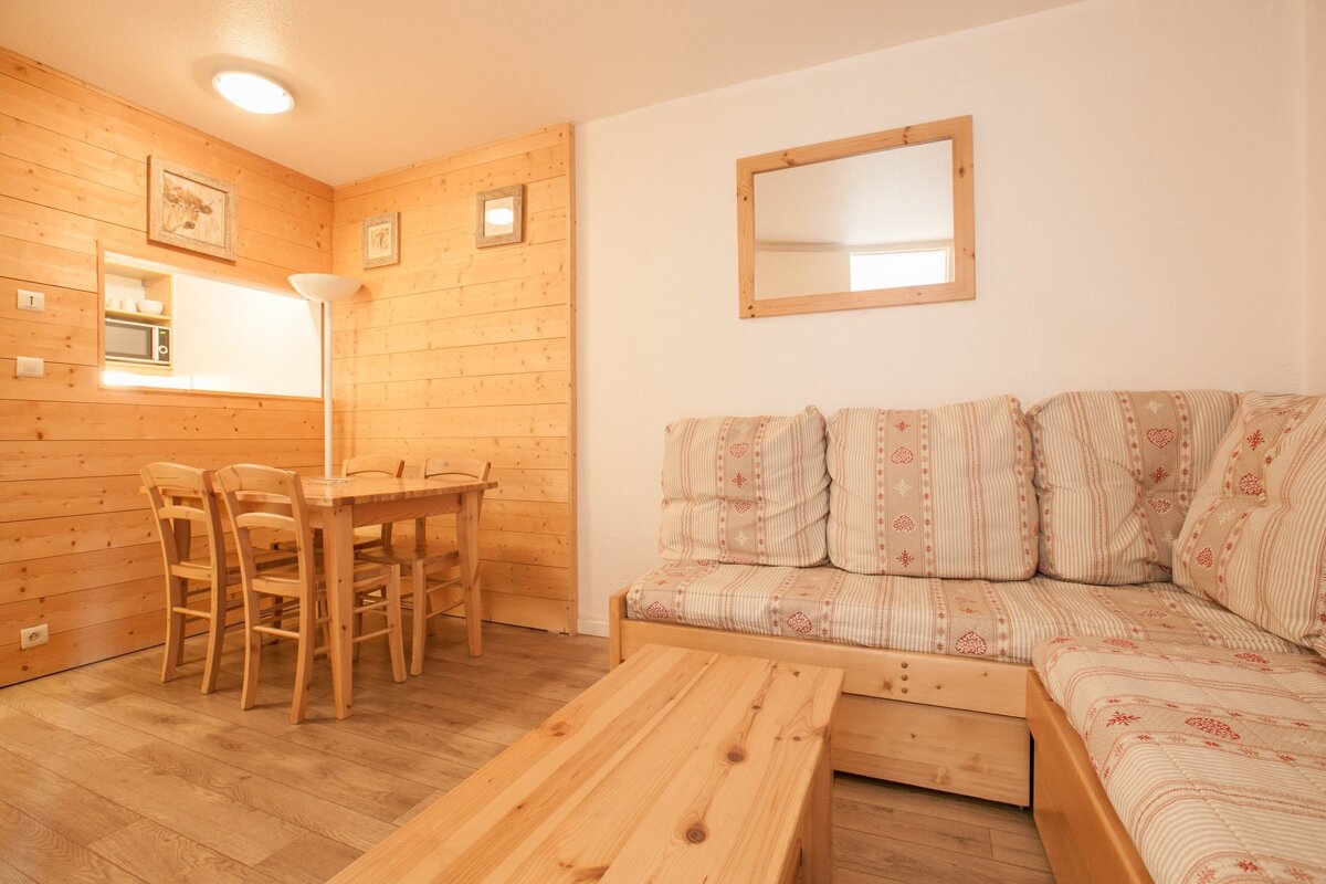 A bright, rustic room with light wood paneling, flooring, and furniture. Features a dining table with chairs, an L-shaped sofa, and a kitchen pass-through.