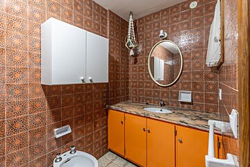 A bathroom featuring brown patterned tiles, an orange vanity with a marble top, white cabinets, an oval mirror, sink, and a bidet.