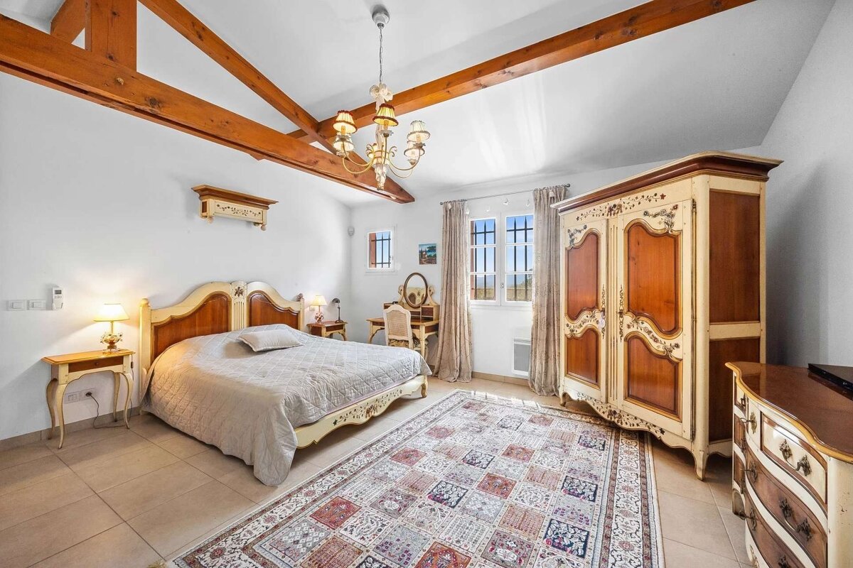 An elegant bedroom with white walls, exposed wooden beams, classic ornate furniture, a large patterned rug, and sunlit windows.