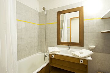 A simple bathroom with a tiled shower/tub, a sink with wooden vanity, mirror, and white towels. Grey tiled walls feature a yellow stripe.