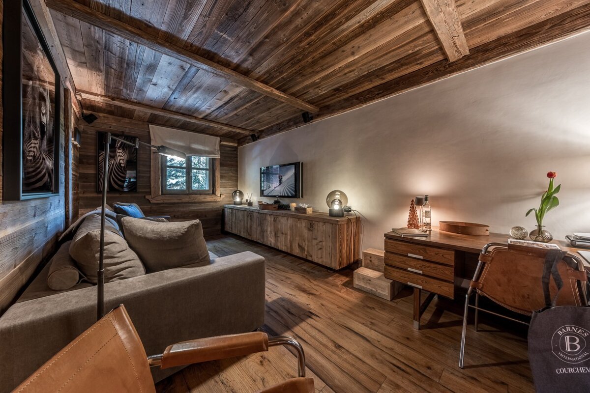 A cozy, wood-paneled living room featuring a comfortable sofa, TV console, and a desk, creating a warm and rustic chalet ambiance.
