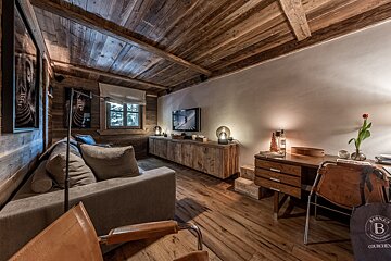 A cozy, rustic chalet-style room with wooden walls, ceiling, and floor. Features a comfy sofa, TV, desk, and warm lighting.