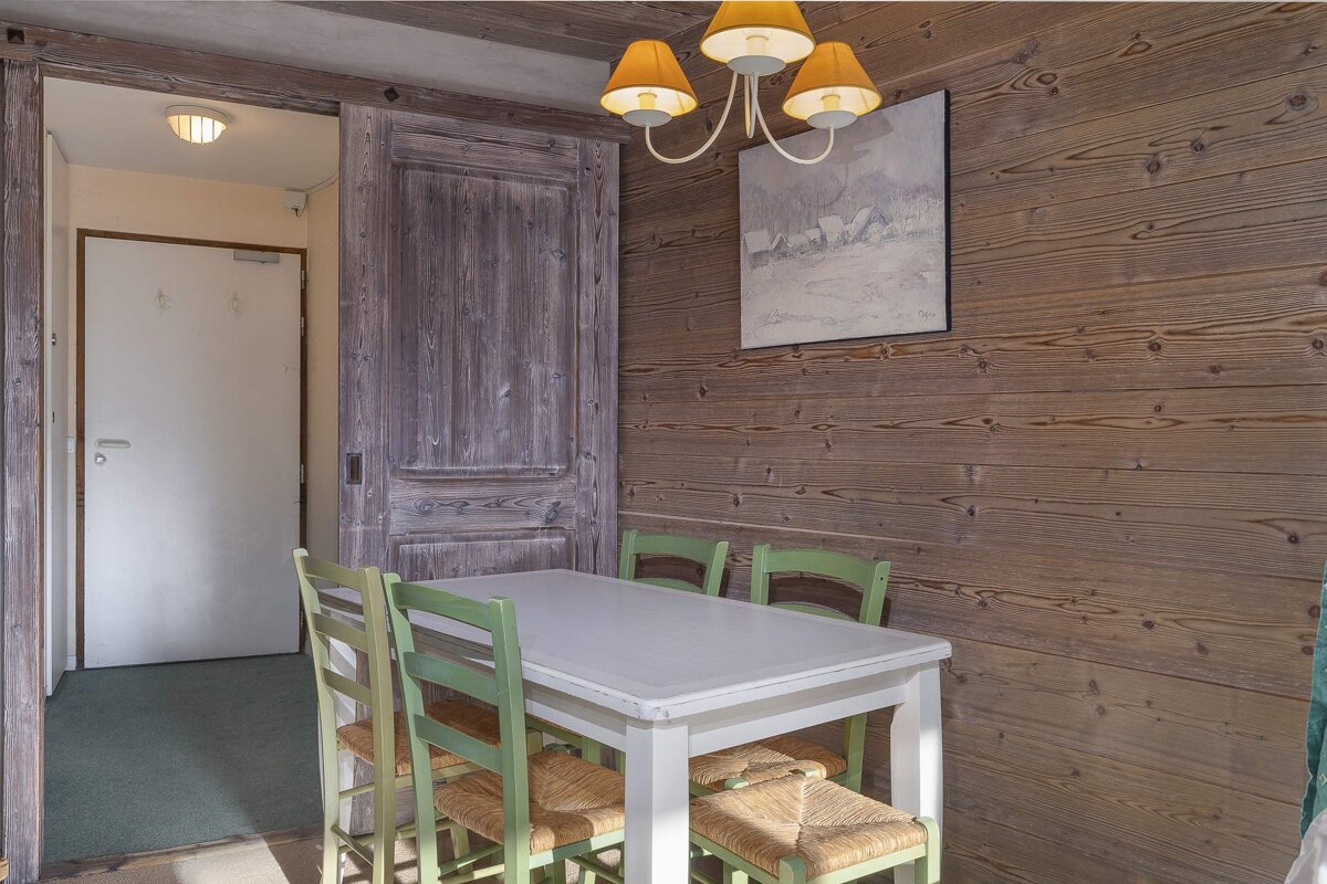 A rustic dining area featuring warm wooden walls, a white table with green chairs, an amber-shaded chandelier, and a landscape painting.
