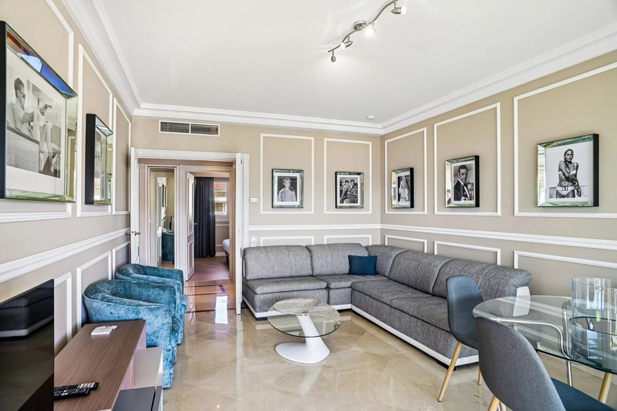 A stylish living room with a large grey L-shaped sofa, glass tables, polished floor, and celebrity photos on light paneled walls.