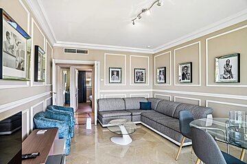 An elegant living room with a grey sectional sofa, blue armchairs, glass tables, and framed black-and-white photos on beige paneled walls.
