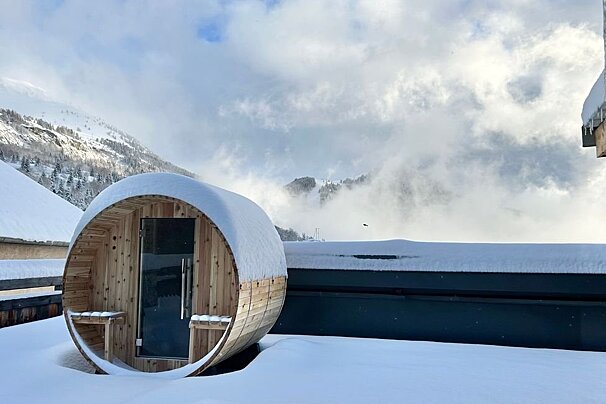 Wooden barrel sauna sits in deep snow on a rooftop overlooking misty winter mountains.