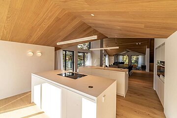 A sleek, modern open-plan kitchen and living space with a dramatic wooden vaulted ceiling, light wooden floors, white islands, and large windows.