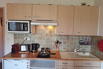 A functional kitchen with light wood cabinetry, a tiled backsplash, and a wooden counter equipped with a microwave, toaster, kettle, stove, and sink.