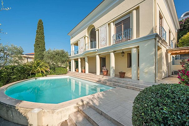 An elegant cream-colored house with columns and balconies overlooks a blue swimming pool, surrounded by lush trees and bushes under a clear sky.