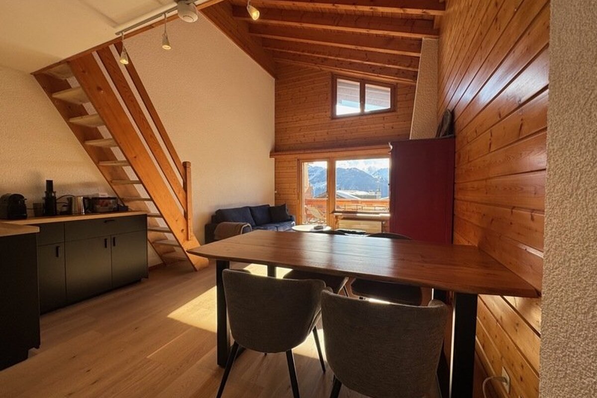 Cozy wooden cabin interior featuring a dining table, staircase, and window view of snowy alpine mountains.