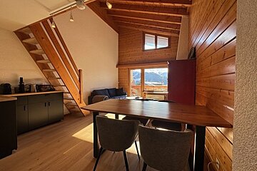 Cozy wooden cabin interior featuring a dining table, staircase, and window view of snowy alpine mountains.