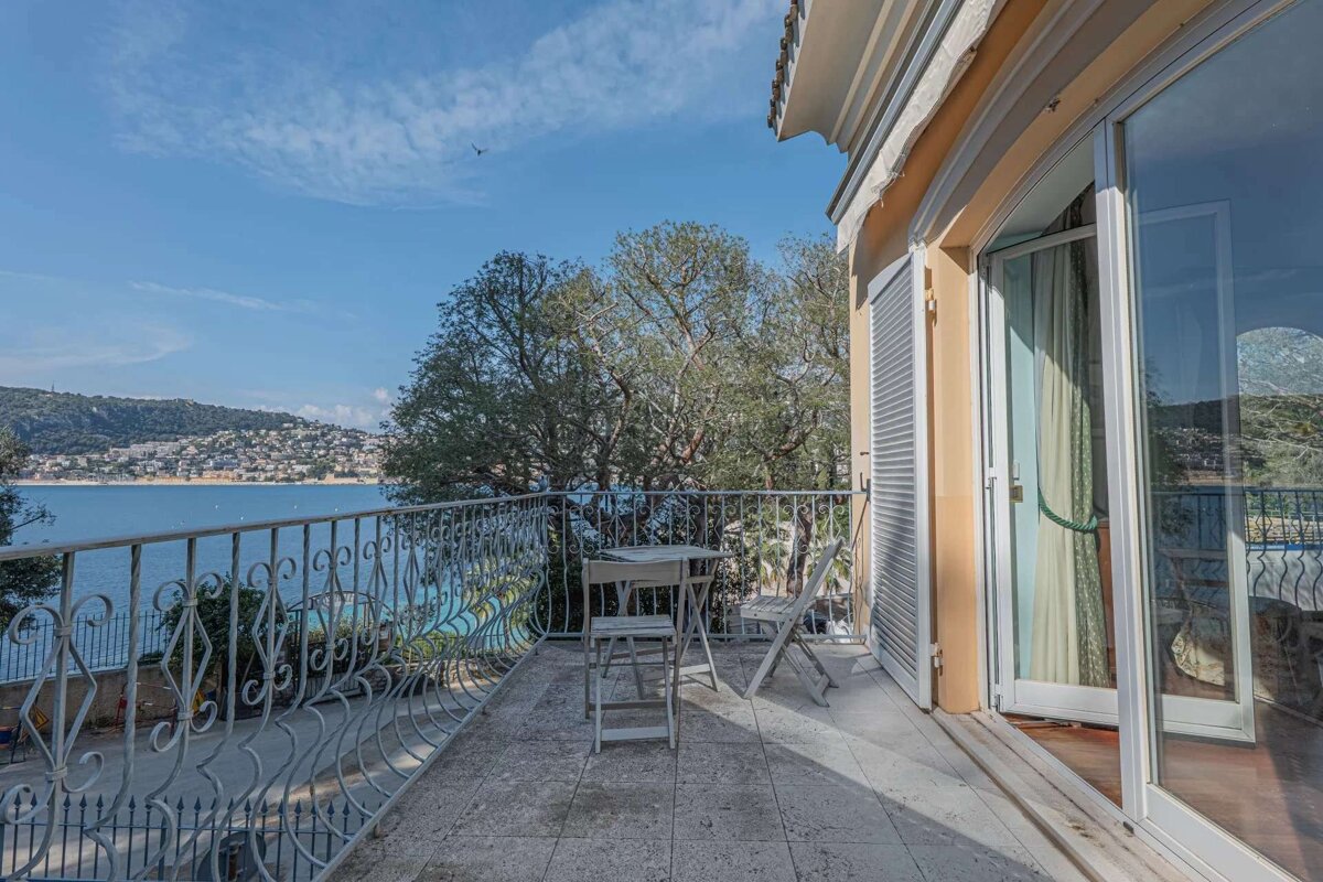 A balcony with a wrought-iron railing overlooks a scenic bay with a coastal town, lush trees, and a clear blue sky. A table and chairs sit on the balcony.