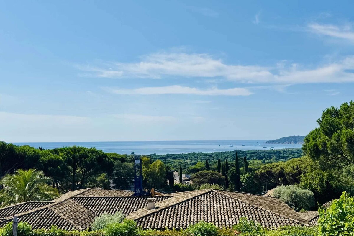 Overlooking red-tiled roofs, a vibrant green landscape slopes towards a calm blue sea, meeting a clear sky with soft clouds and distant land.