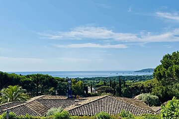 A panoramic view overlooking terracotta rooftops, lush green trees, the sparkling blue sea, and a clear sky with scattered clouds.