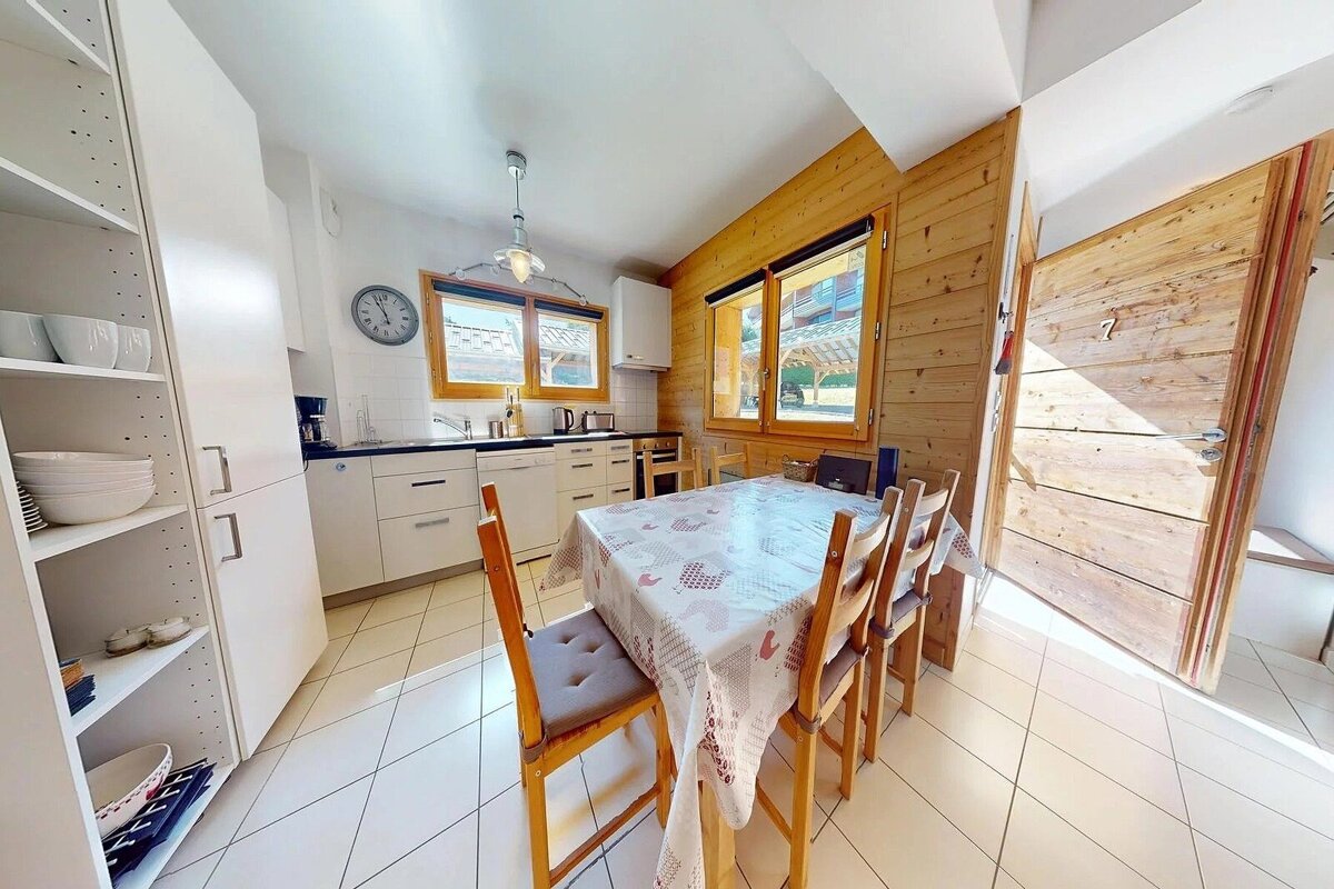 A cozy kitchen and dining area features white cabinets, a table with a patterned cloth, wooden chairs, wood-paneled walls, and a wooden door.