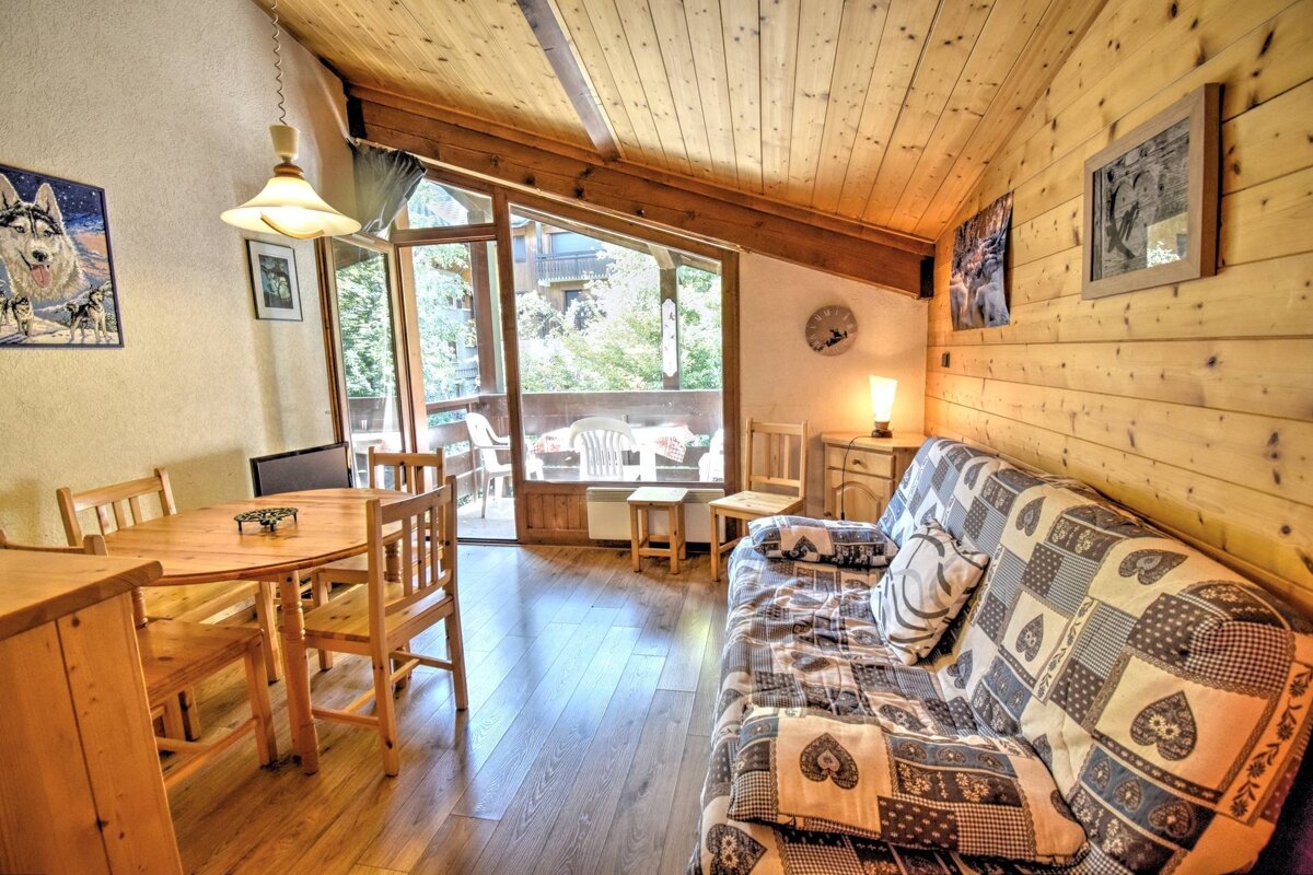 A rustic, wood-paneled living room features a patterned sofa, a wooden dining table, and large glass doors opening to a green balcony.