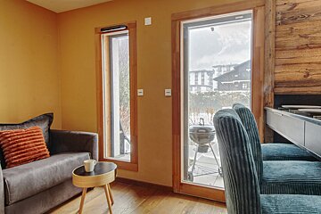 A cozy room with warm walls, wooden floors, and a sofa, featuring two large windows looking out to a snowy village and a grill on a patio.