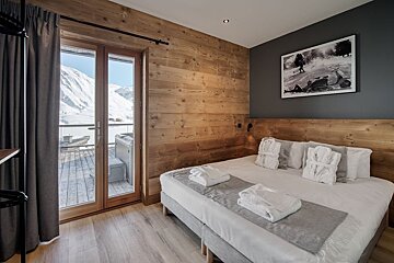 Cozy bedroom with wooden walls, a bed with white linens, and a balcony overlooking snowy mountains, featuring a hot tub and dark grey accent wall.