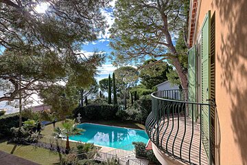 A sunny view from a curved balcony, overlooking a bright blue pool, lush green gardens, and the sea in the distance, framed by trees.