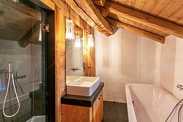 A rustic attic bathroom features exposed wooden beams, a glass shower, modern vanity with vessel sink, sleek bathtub, and pendant lights.