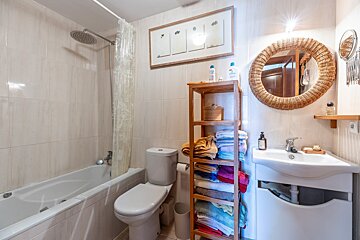 A bright, light-tiled bathroom with a bathtub, shower, toilet, and sink. A round wicker mirror and a wooden shelf with colorful towels add warmth.