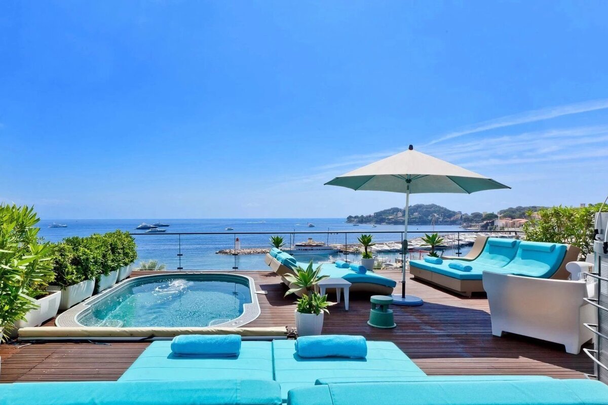 Luxury rooftop terrace with a hot tub, turquoise loungers, and umbrella, overlooking a vibrant blue sea and a harbor full of boats.