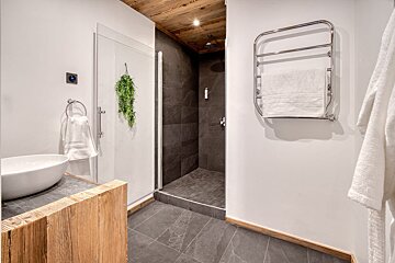 A modern bathroom with a dark tiled shower, white walls, wooden ceiling, white basin, and a towel warmer.