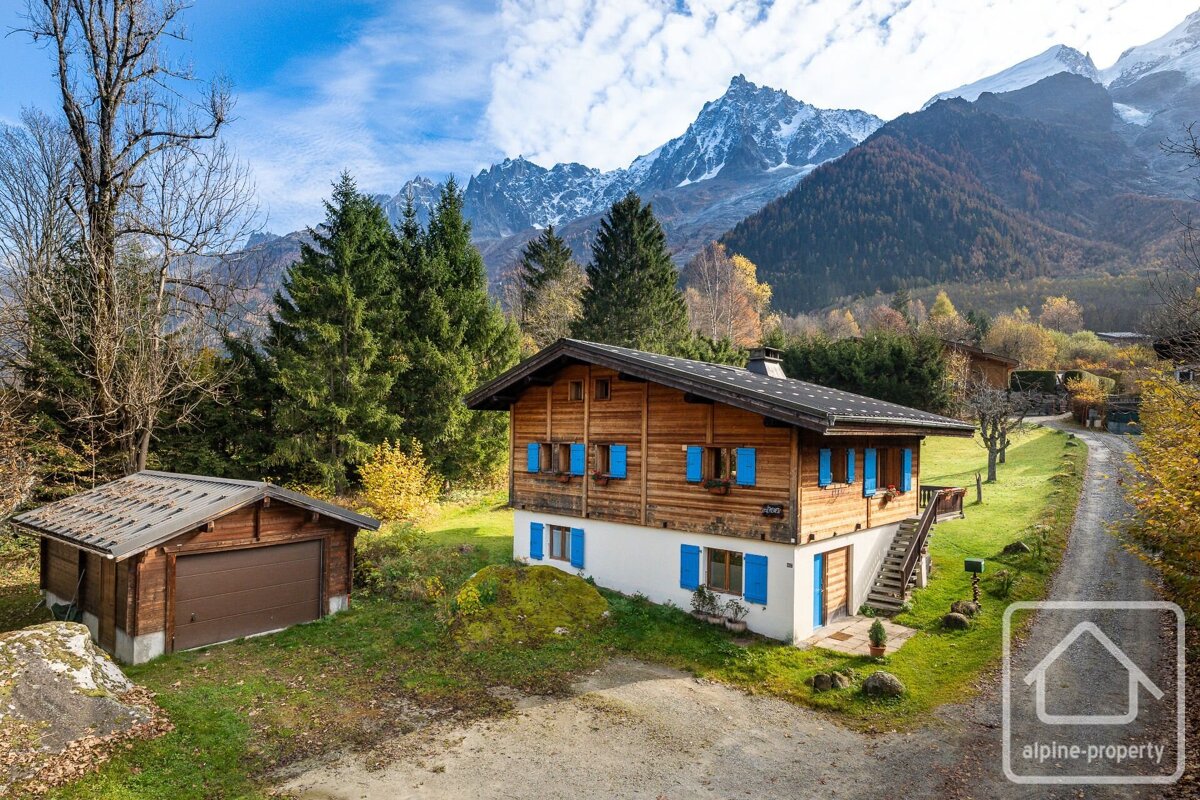 A picturesque wooden chalet with blue shutters and a garage set against towering snow-capped mountains and colorful autumn trees on a sunny day.