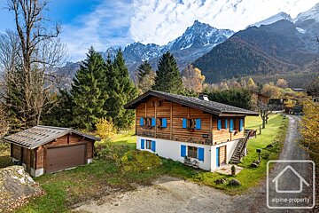 A picturesque wooden chalet with blue shutters and a garage set against towering snow-capped mountains and colorful autumn trees on a sunny day.