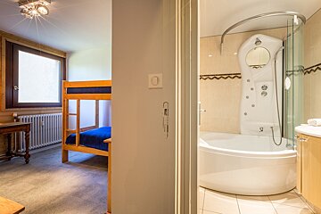 A bedroom with wooden bunk beds and a window is visible through an opening to a modern bathroom with a white tub and shower unit.