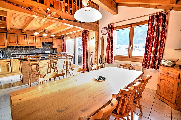 Chalet-style open-plan kitchen and dining room with wooden interiors, exposed beams, a large table, and windows showing a snowy view.