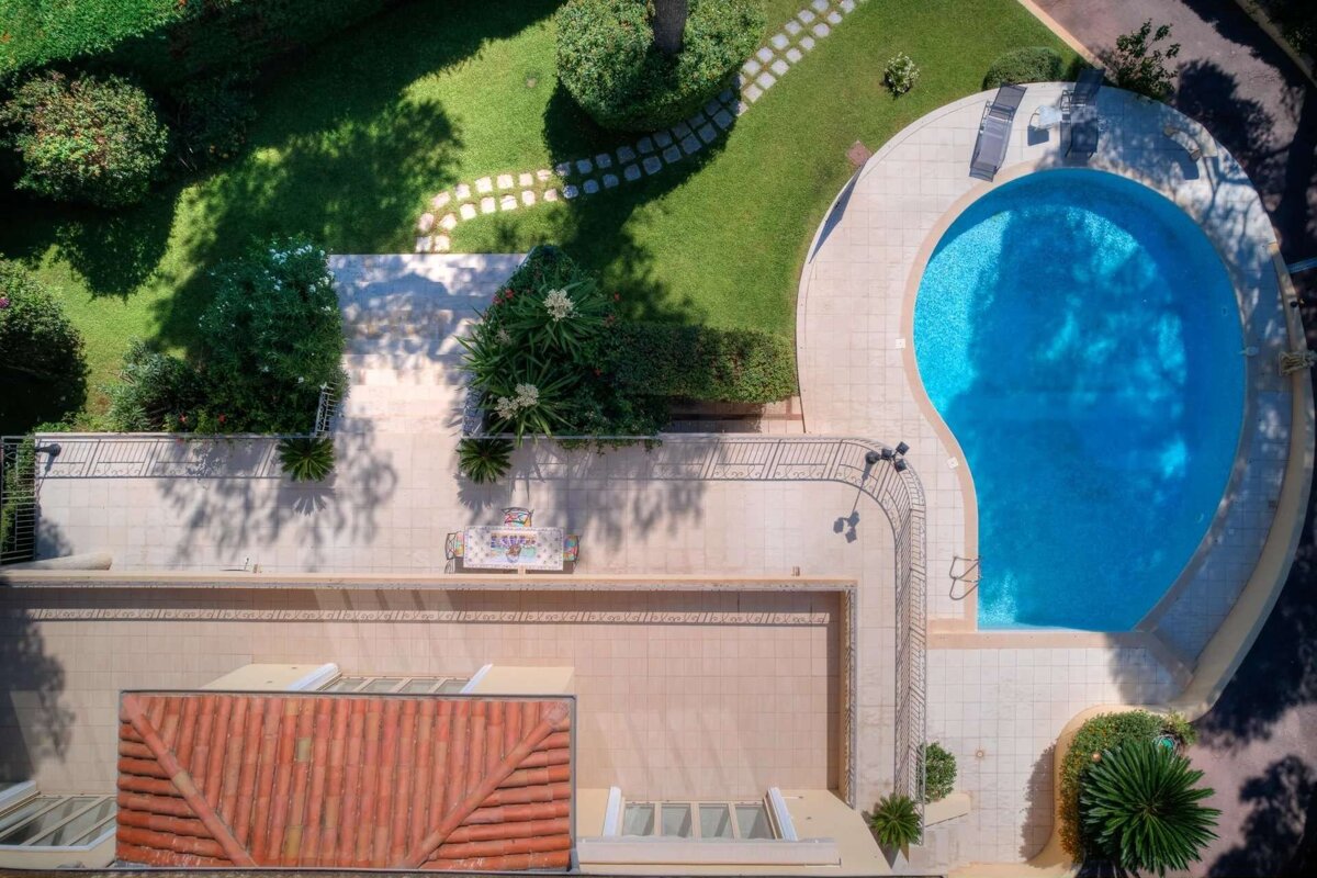 Aerial view of a property featuring a bright blue swimming pool, paved patio, lush green garden with a stone path, and a red-tiled rooftop.