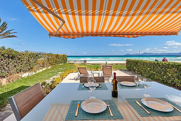 Outdoor dining table under a striped awning, overlooking a sunny beach with turquoise water and distant mountains.