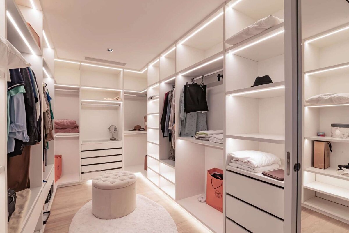 A modern, bright white walk-in closet with integrated LED lighting, organized clothes on shelves and racks, plus a central tufted ottoman.
