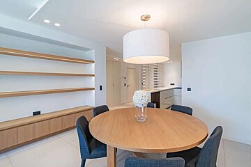 Modern dining area with a round wooden table, dark chairs, white chandelier, and illuminated built-in shelves. A bright kitchen is seen beyond.