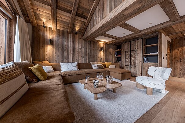 Cozy, rustic chalet living room with wood paneled walls, a large brown sectional sofa, wooden coffee tables, and a modern white armchair.