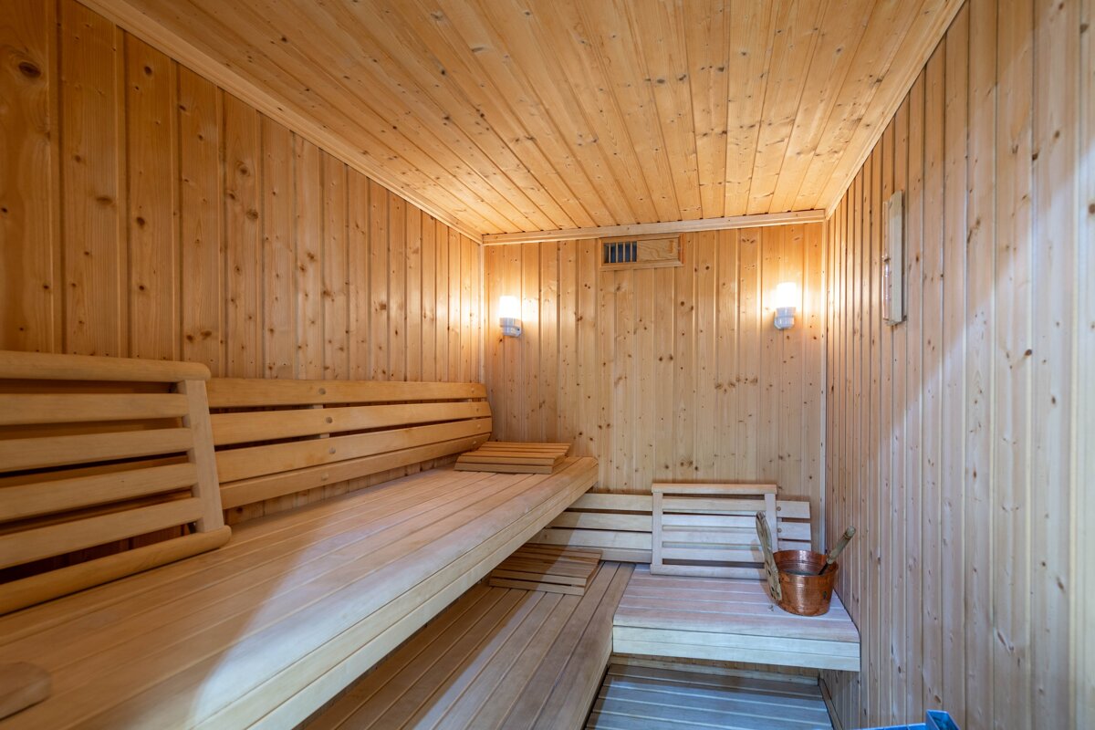 A bright, empty wooden sauna with multi-level benches, warm wall lighting, and a copper bucket and scoop, ready for relaxation.