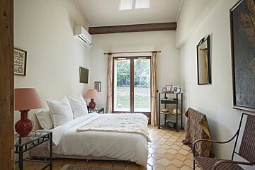 A serene bedroom with a white bed, patterned tile floor, large double doors looking out to a garden, and warm, rustic accents.