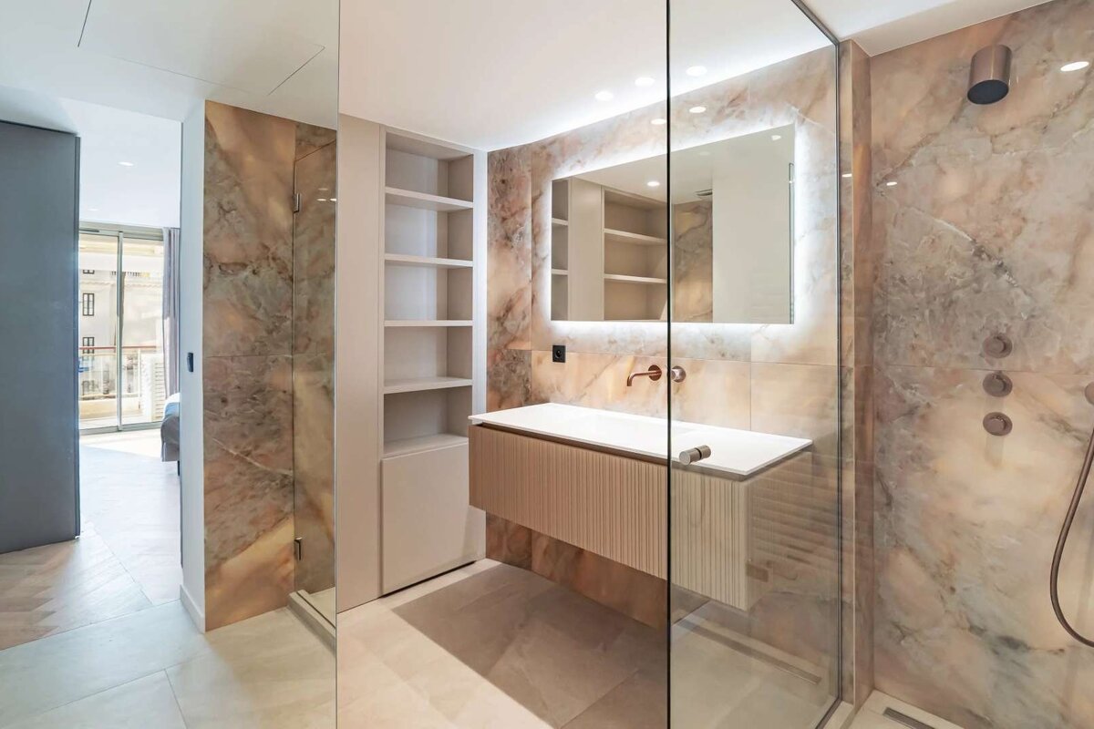 Modern bathroom with warm marble walls, a floating vanity, backlit mirror, and glass shower. Features neutral tones and built-in shelving.