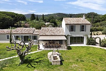 A charming house with terracotta roofs and grey shutters sits on a lush green lawn. An outdoor dining area, an old tree, and a wooded hillside are visible.