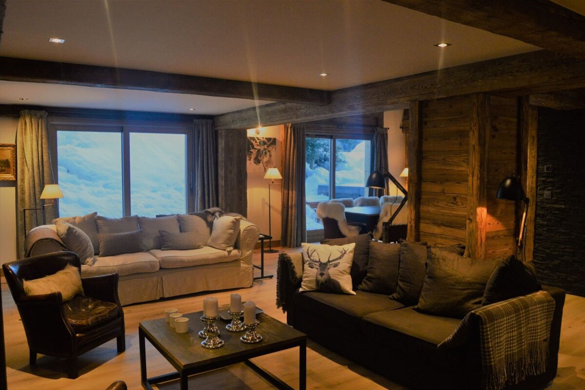 Cozy chalet living room with two sofas, wood accents, and warm lighting. Large windows reveal a snowy mountain view, perfect for relaxation.
