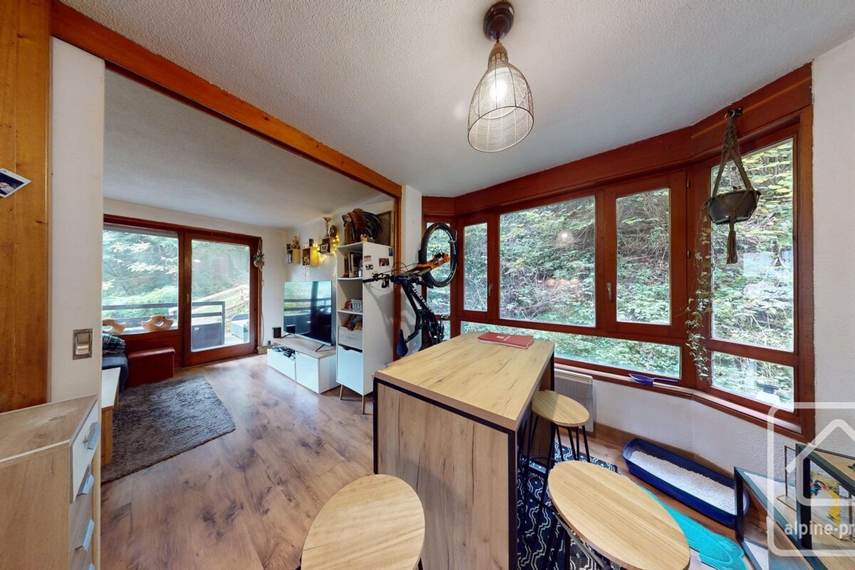 A cozy apartment dining space with a high table, stools, wooden floors, large windows overlooking trees, and an adjoining living area.