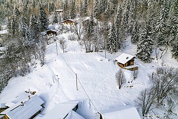 Aerial view of a snow-covered mountain landscape with traditional wooden cabins nestled among dense pine trees. Footprints crisscross the white slopes.
