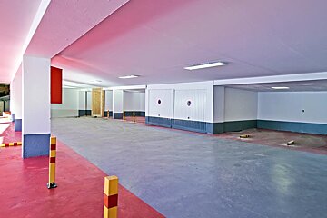 Empty indoor parking garage with a pinkish ceiling, red and grey floor, white pillars, and closed garage doors.