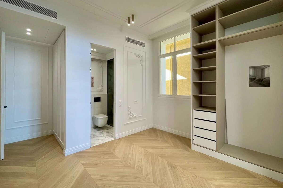 A clean, bright room featuring light wood herringbone flooring, white walls, built-in shelves, a window, and an adjacent modern bathroom.