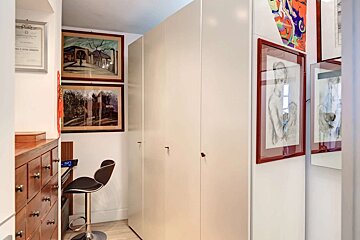 A narrow room with white built-in cabinets, a wooden drawer chest, and various framed artworks. A black bar stool sits by a small desk.