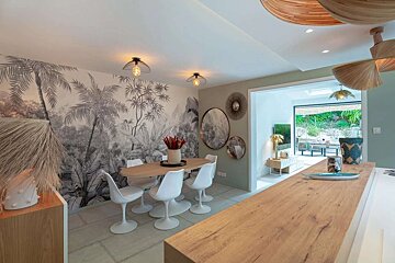 A chic dining room featuring grayscale tropical palm tree wallpaper, an oval wood table with white chairs, and an open view to a lush outdoor area.