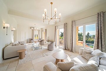 Bright, spacious living room with cream furniture, tile floors, a patterned rug, gold chandeliers, and large windows.