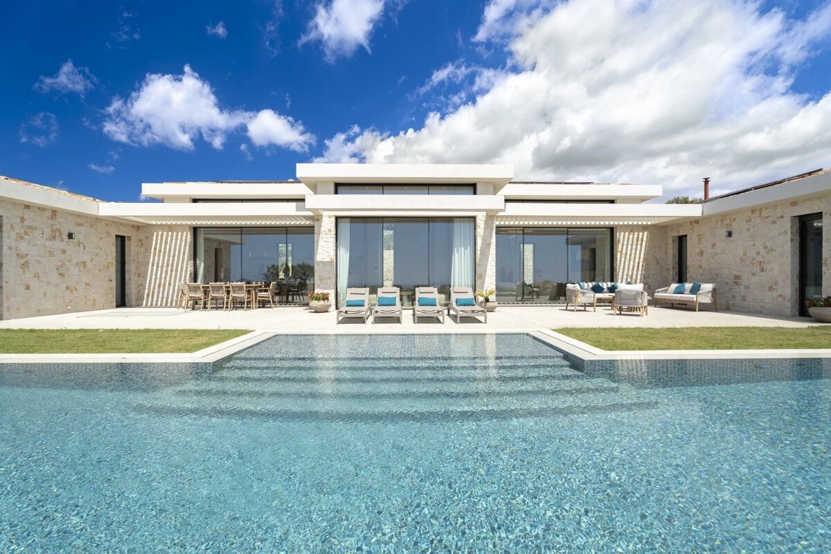 A modern luxury villa with stone walls, floor-to-ceiling glass, a spacious patio, outdoor dining & lounging, and a large pool under a bright blue sky.