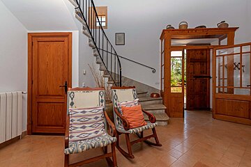 Rustic entryway with stone spiral stairs, two wooden rocking chairs, terracotta floor, and wooden doors leading to a lush garden.